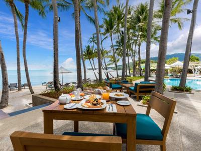 Ein gedeckter Tisch mit Essen am Strand unter Palmen neben einem Pool mit Meerblick.