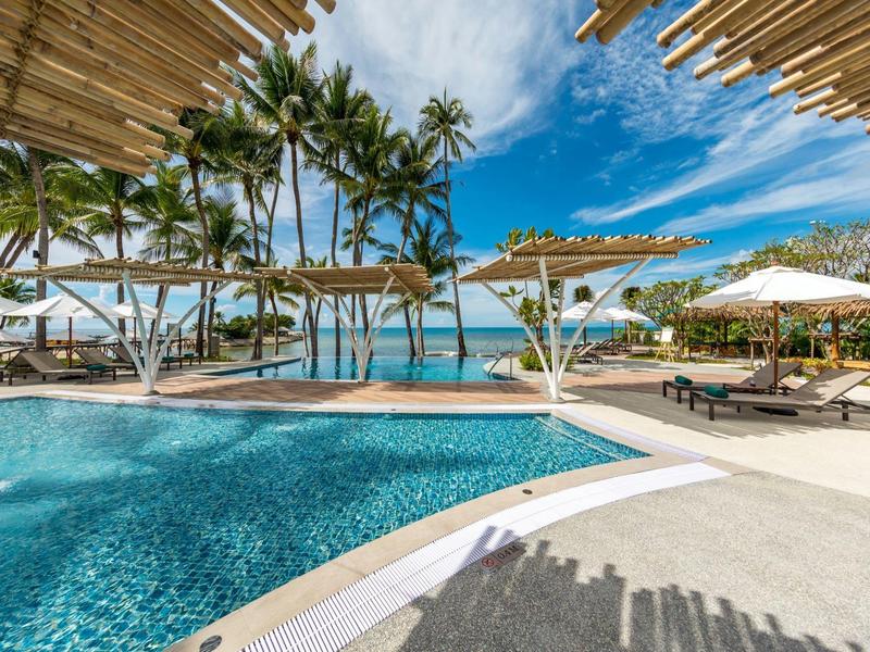 Luxusní bazénová oblast s palmami a bílými slunečníky u moře pod jasnou oblohou.