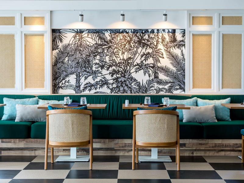 Elegantní hotelová lounge s zelenou čalouněnou lavicí, dřevěnými stoly a černobílým obrazem na stěně.