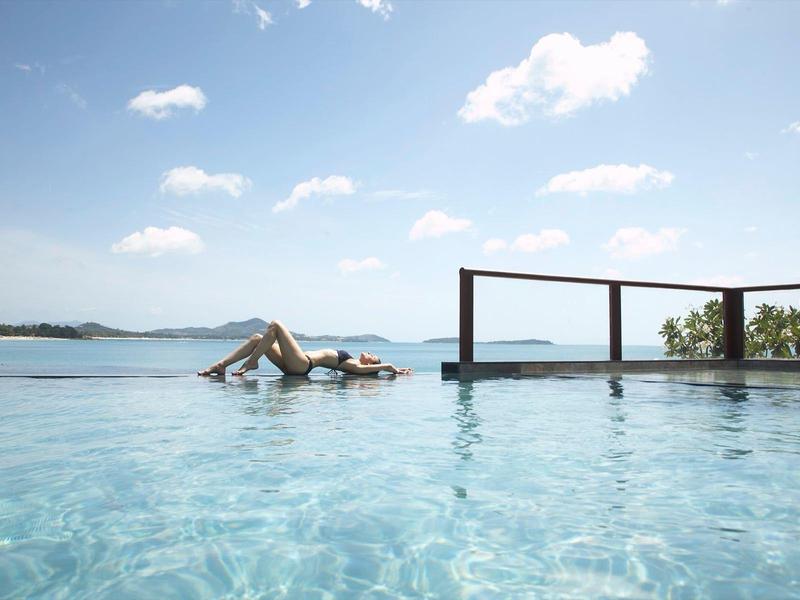 Femme se détendant dans une piscine à débordement avec vue sur la mer et ciel bleu.