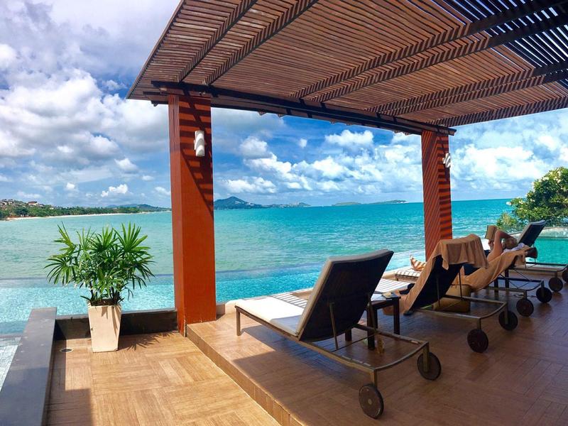 Chaises longues sous une tonnelle en bois avec vue sur la mer, ciel bleu et palmiers.