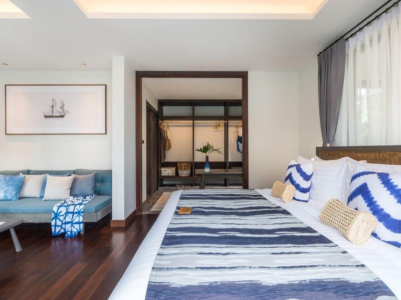 Modern eingerichtetes Schlafzimmer mit blau-weißer Bettwäsche und Sofa neben einem Fenster mit Vorhängen.