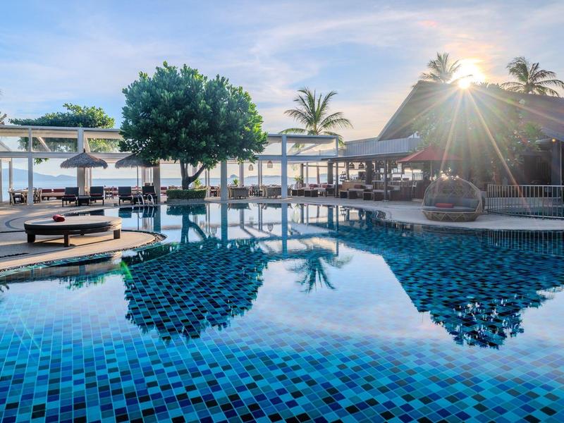 Hotelpool mit Sonnenliegen, Palmen und Blick auf das Meer bei Sonnenuntergang.