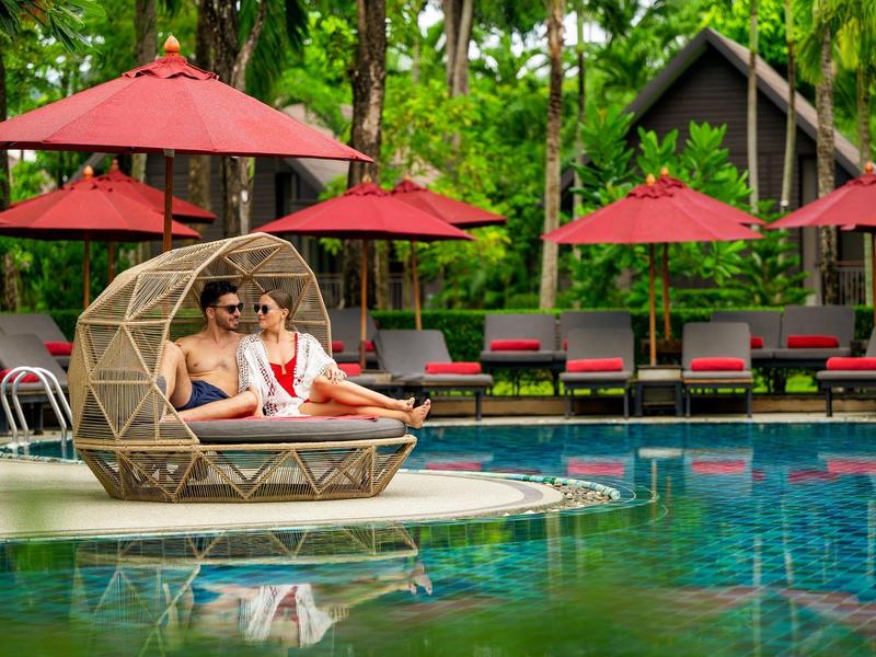 Paar entspannt in einem runden Lounge-Sessel am Pool mit roten Sonnenschirmen und grüner Hotelanlage.