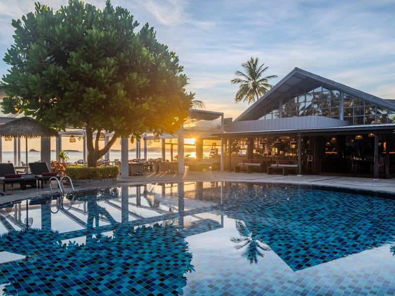Blick auf einen Pool mit Sonnenuntergang, umgeben von Bäumen und einem Hotelgebäude.