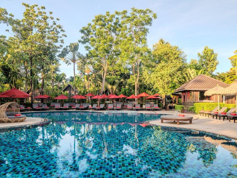 Großer Pool mit Liegestühlen und Sonnenschirmen, umgeben von Bäumen und Gebäuden.