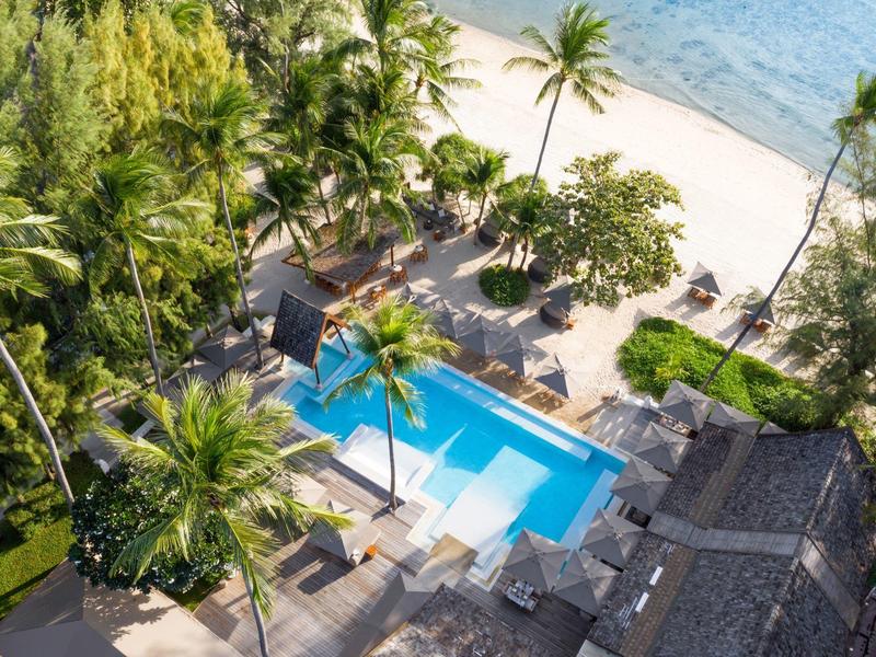 Hotel de lujo con piscina y palmeras junto a la playa de arena blanca y mar azul.