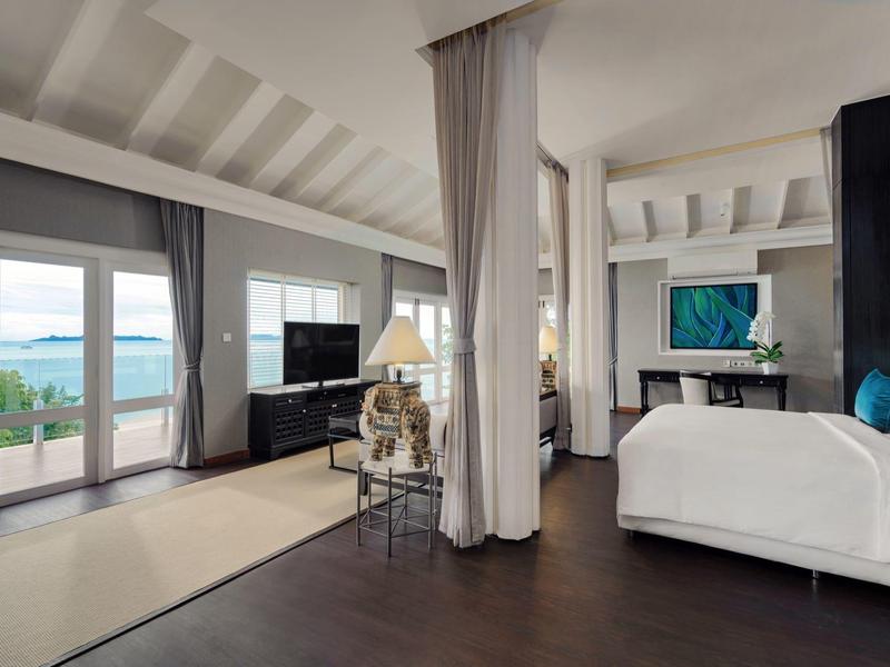 Habitación de hotel luminosa con cama grande, zona de estar y vista al mar a través de grandes ventanales.