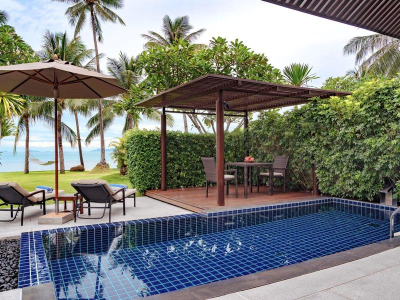 Piscina al aire libre moderna con tumbonas y sombrillas, rodeada de vegetación tropical con vista a la playa.