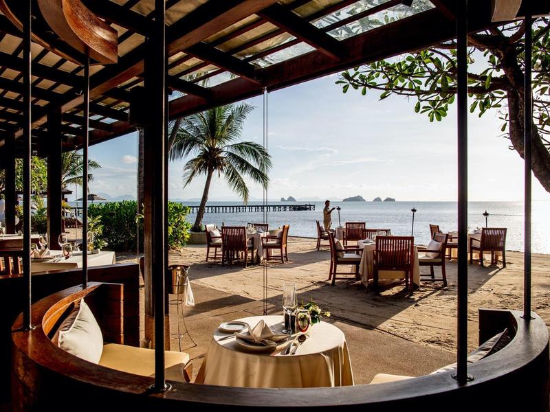 Área de restaurante abierta con vista al mar, mesas con manteles blancos y palmeras al fondo.