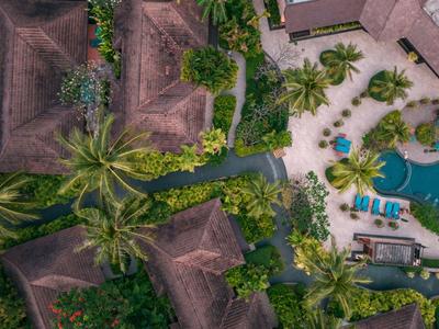 Luftaufnahme eines tropischen Resorts mit Pool, Palmen und mehreren Gebäuden mit braunen Dächern.