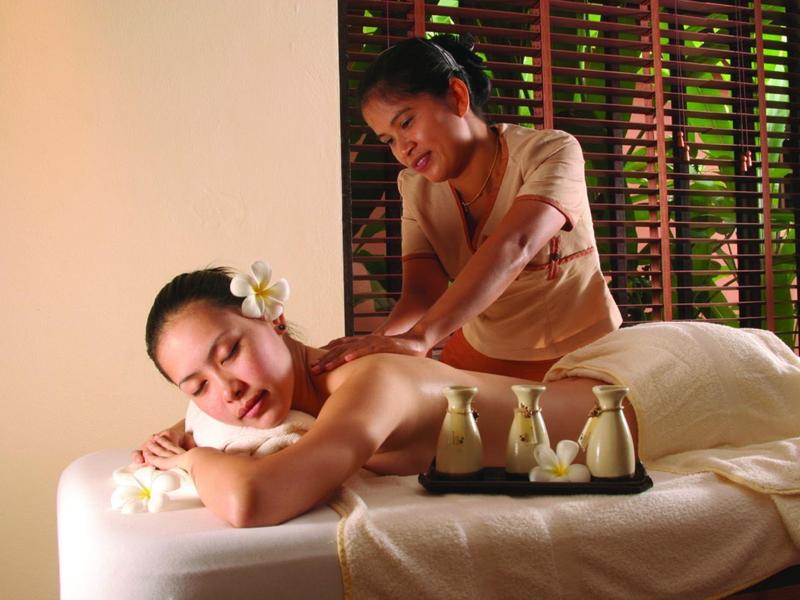 Mujer recibiendo masaje relajante en mesa con flor en el cabello y aceites de aromaterapia.