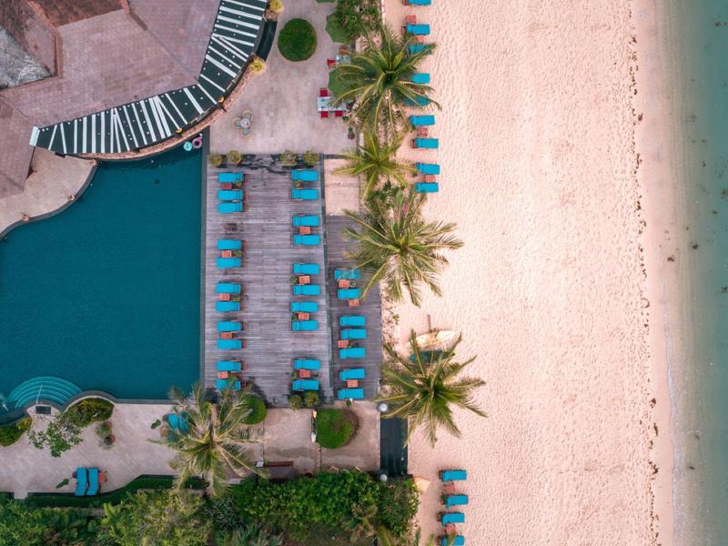 Vista aérea de una piscina junto a una playa de arena con tumbonas y palmeras