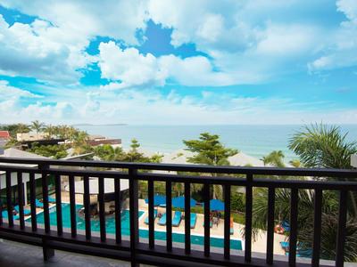 Balcón con barandilla negra con vista a la piscina, árboles y mar bajo un cielo azul con nubes