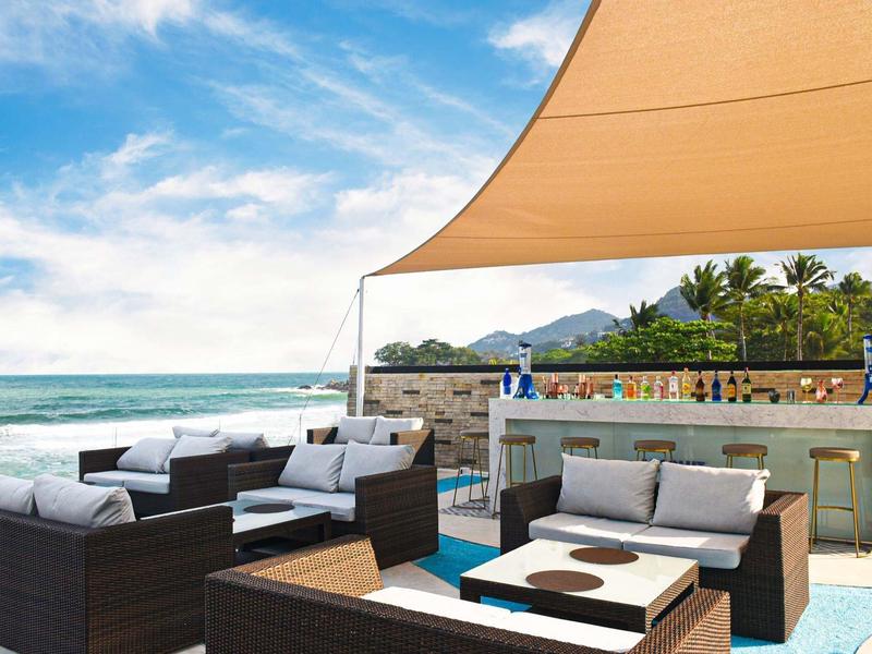 Área exterior moderna con sofás, toldo y bar con vistas al mar y montañas al fondo.