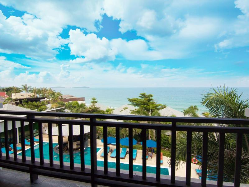 Balcón con barandilla negra con vista a la piscina, árboles y mar bajo un cielo azul con nubes