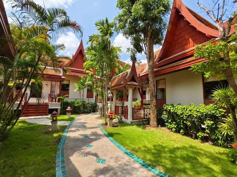 Un chemin de jardin soigné mène à des bâtiments thaïlandais traditionnels aux toits rouges et à la végétation tropicale.
