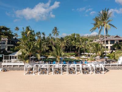 Hotel con piscina e lettini su spiaggia sabbiosa, circondato da palme sotto cielo blu.