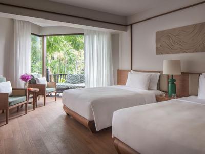 Camera d'hotel con due letti, una sedia e un tavolo vicino a una grande finestra con vista su piante verdi.