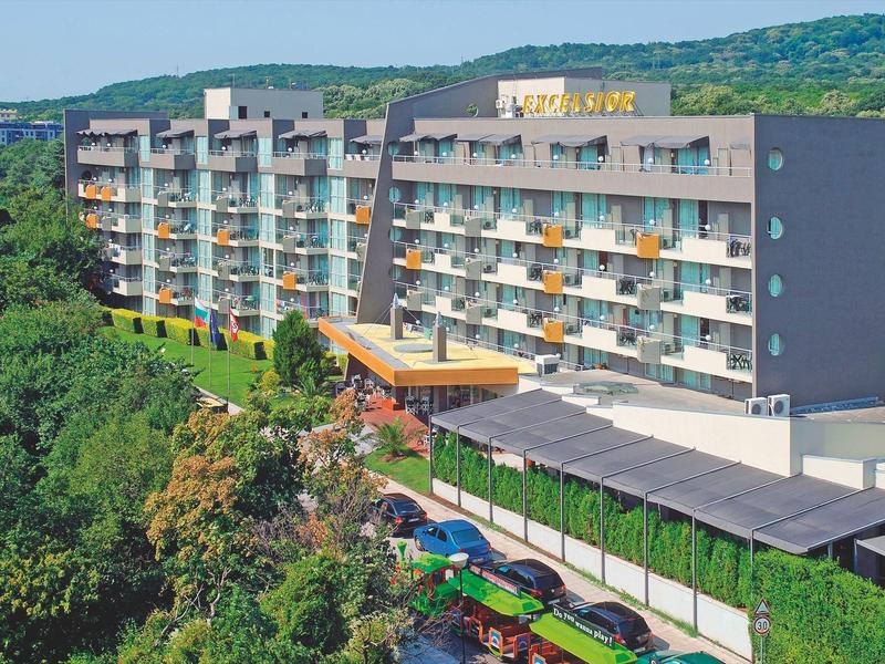 Groot meerlagig hotelgebouw met balkons, omgeven door groen landschap en heuvels.