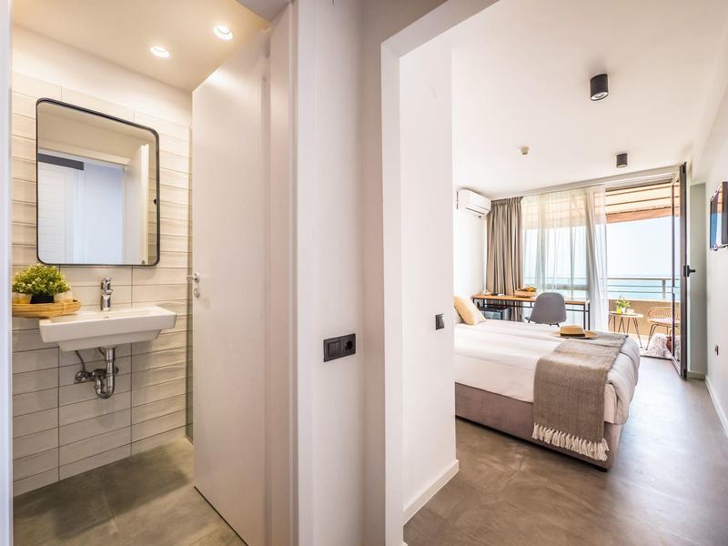 Lichte hotelkamer met groot bed en aangrenzende badkamer met wastafel en spiegel.