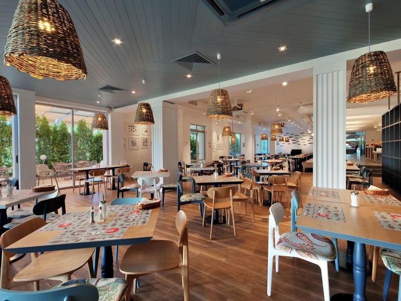 Modern restaurant met houten tafels, stoelen en grote ramen met uitzicht op groen.