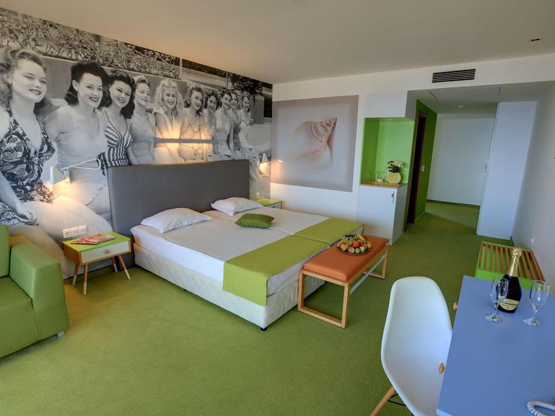 Modern hotelkamer met tweepersoonsbed, groen tapijt, bank, bureau en fotobehang aan de muur.