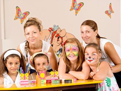 Zwei Frauen und drei Mädchen mit bemalten Gesichtern sitzen an einem Tisch mit Spielzeug, Schmetterlinge an der Wand.