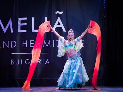 Zwei Tänzer in farbenfrohen Kostümen mit langen roten Bändern auf einer Bühne.