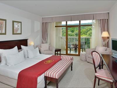 Helles Hotelzimmer mit Doppelbett, Schreibtisch, Fernseher und Balkon mit Blick ins Grüne.