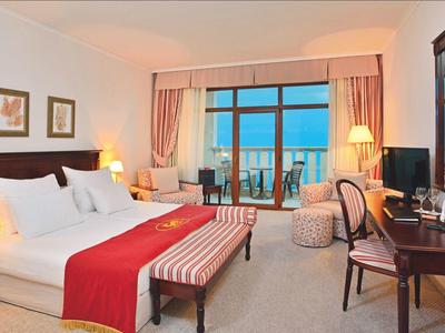 Heller Hotelzimmer mit großem Bett, rot-weißem Bettläufer und Balkon mit Blick auf das Meer.