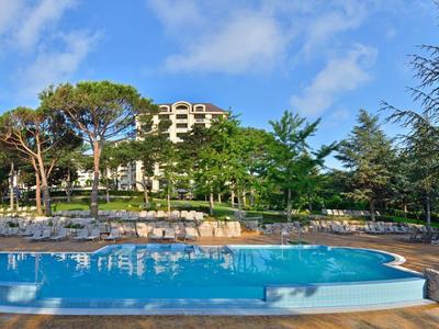 Großer Pool mit blauer Wasserfläche, umgeben von Stühlen und grünen Bäumen unter blauem Himmel.