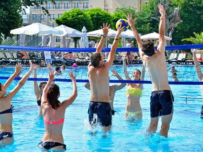 Menschen spielen Volleyball im Schwimmbecken eines Hotels unter weißen Sonnenschirmen.