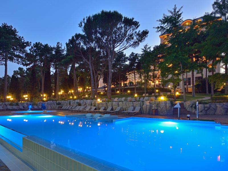 Beleuchteter Außenpool vor einem Hotelgebäude am Abend mit Sitzgelegenheiten.