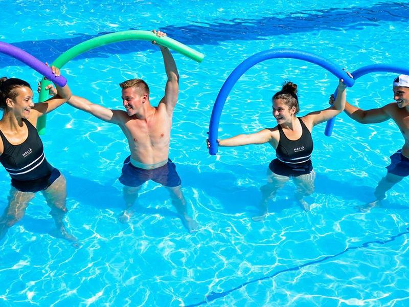 Vier Personen mit Schwimmnudeln spielen im blauen Pool Wasser.