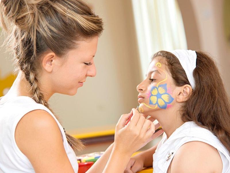 Frau malt ein farbenfrohes Schmetterlingsgesicht auf Mädchen im hellen Innenraum.