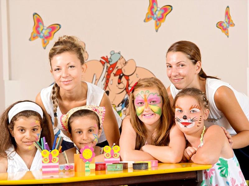 Zwei Frauen und drei Mädchen mit bemalten Gesichtern sitzen an einem Tisch mit Spielzeug, Schmetterlinge an der Wand.