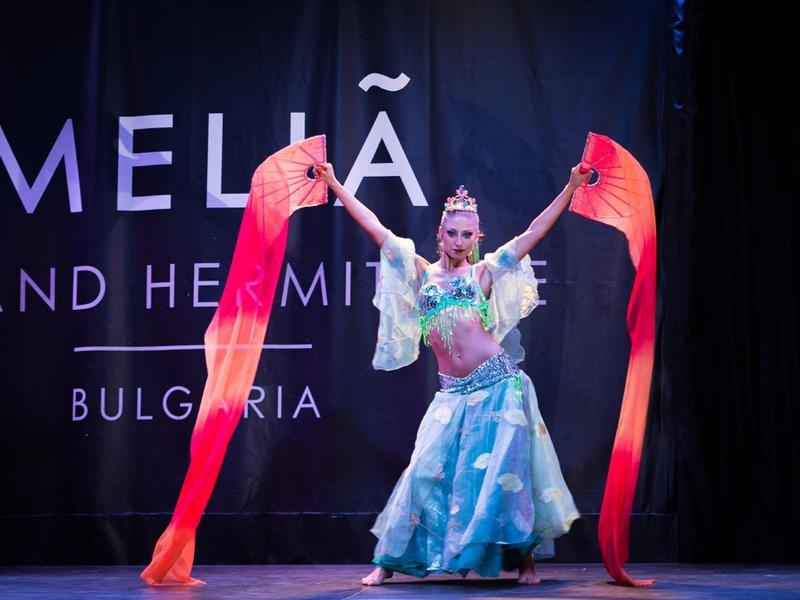 Tänzerin auf Bühne mit blauem Rock und langen roten Schleifen, Hintergrund schwarzer Vorhang mit Text.