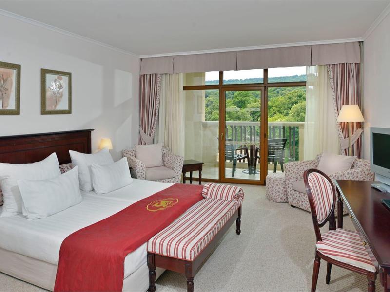 Helles Hotelzimmer mit Doppelbett, Schreibtisch, Fernseher und Balkon mit Blick ins Grüne.