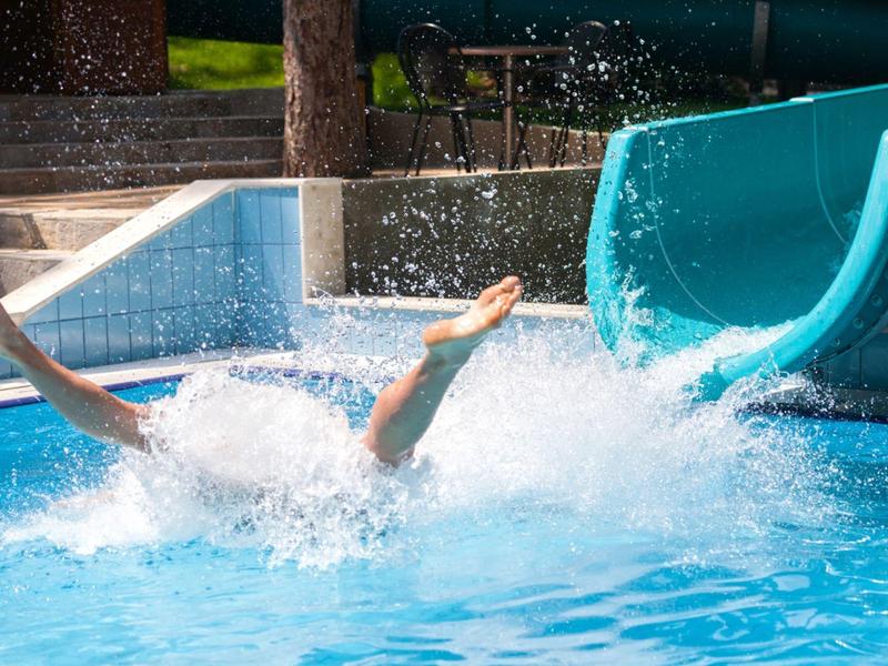 Eine Person rutscht mit Armen in die Luft aus einer blauen Wasserrutsche in einen Pool.