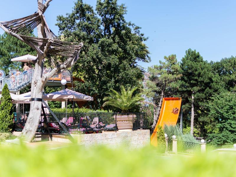 Spielplatz mit Holzgerüst und orangefarbener Rutsche in einer grünen Parkumgebung bei Sonnenschein.