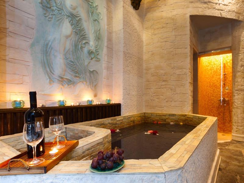 Luxuriöses Bad mit Marmorwanne, Weinflasche, Gläsern und warmem Kerzenlicht in hellem Steinraum.