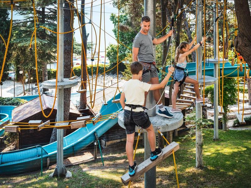 Drei Personen erklimmen einen Hochseilgarten im Freien mit Bäumen und Kletterelementen.
