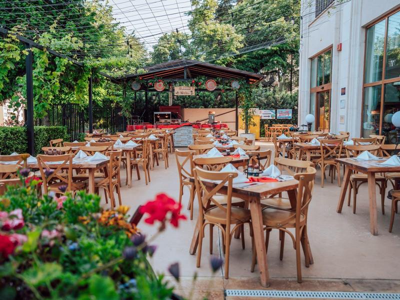 Offenes Restaurant mit Holzstühlen und Tischen unter einem Glasdach, umgeben von Pflanzen.