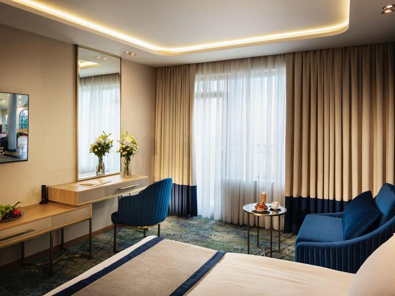 Modern eingerichtetes Hotelzimmer mit dunkelblauen Sesseln, Holzschreibtisch und großen Vorhängen.