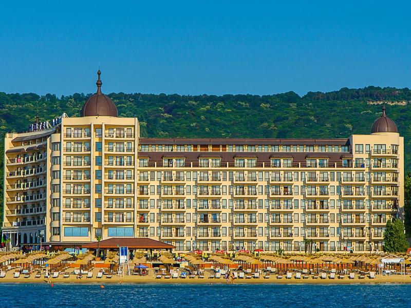 Großes mehrstöckiges Hotel mit vielen Balkonen, davor Sandstrand und blaue See, im Hintergrund grüne Hügel.