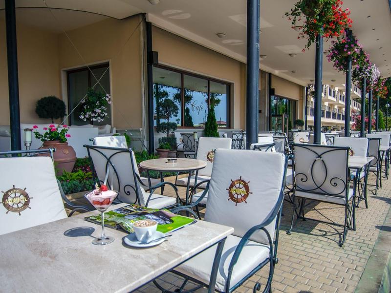 Straßencafé mit weißen Metallstühlen und Tischen, blühenden Hängepflanzen und Sonnenschutzdach.