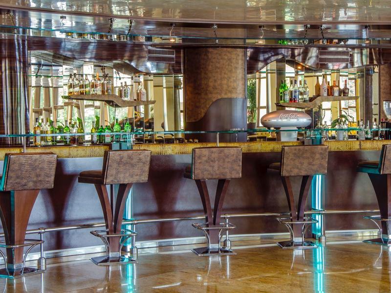 Elegante Bar mit fünf hohen Holzstühlen, Spiegeln und beleuchteten Flaschenregalen im Hintergrund.