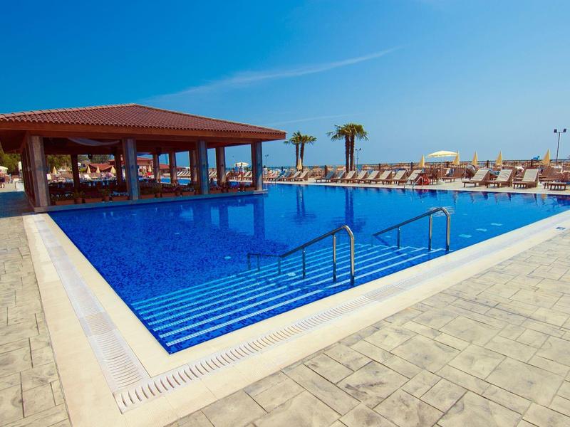 Ein großer blauer Pool mit Treppen, umgeben von Liegestühlen und einer überdachten Bar bei sonnigem Himmel.