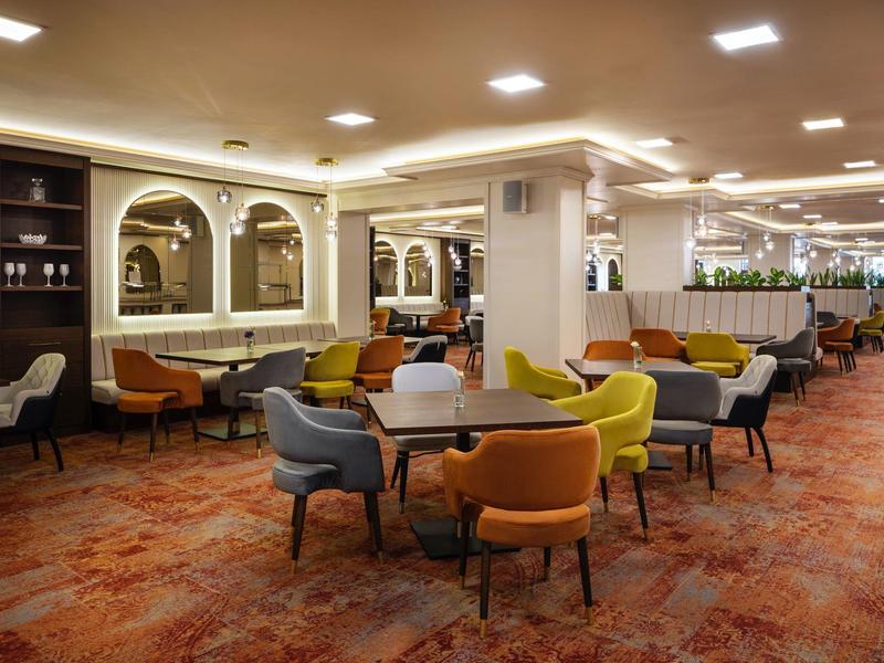 Großer, heller Raum mit bunten Stühlen und runden Tischen auf gemustertem Teppichboden.